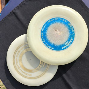 May include: Two white plastic frisbees. The top frisbee has blue text that reads "World Class Night Lighter 133 G Model Frisbee". The bottom frisbee has gold text that reads "Moonlighter 110 G Model Frisbee".