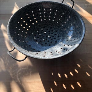 May include: A speckled, dark gray colander with numerous small holes and two black handles. The kitchenware is made of metal and sits on a wooden surface. The colander is designed for draining liquids from food items.