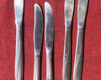 May include: Five stainless steel butter knives with starburst designs on the handles, arranged on a red cloth. The knives have a curved blade and a decorative handle. The red cloth provides a contrasting background.