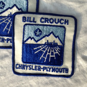 May include: Two embroidered patches with a white background and blue trim. The patches feature the text "BILL CROUCH" and "CHRYSLER-PLYMOUTH". The design includes a mountain range and a star.