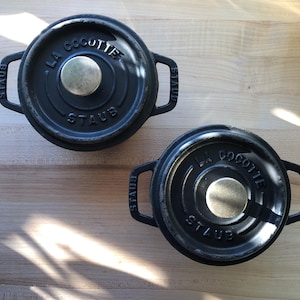 May include: Two black cast iron Staub La Cocotte Dutch ovens with lids. Each lid has a brass-colored knob and the brand name is embossed on the lid. The ovens are on a light wood surface.