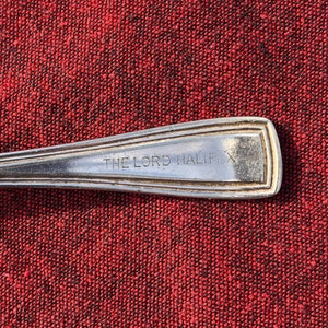 May include: Close-up of a silver spoon with the engraved text "THE LORD HALIFAX". The spoon is set against a red textured background, highlighting the details of the silverware.