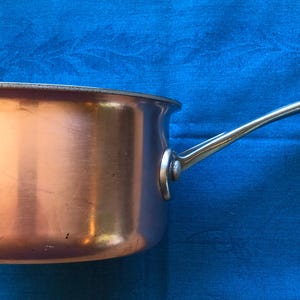 May include: A copper saucepan with a long, curved, silver-colored handle. The pot has a rounded body and a slightly flared rim. The copper surface reflects light, and the handle is attached with a rivet. The background is a solid blue color.