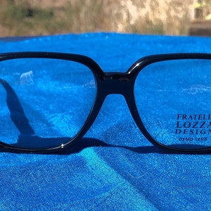 May include: Black square-shaped eyeglasses with blue tinted lenses. The brand name "Fratelli Lozza Design" is printed on the right lens in blue.