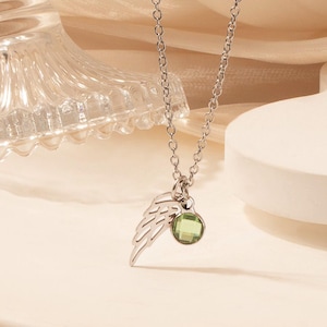 May include: A silver necklace with a delicate chain, featuring a small wing charm and a round, green gemstone pendant. The necklace is displayed on a white, curved surface, with a glass jar in the background.