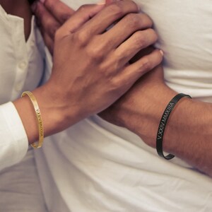 Op de afbeelding: Een stel draagt bijpassende gouden en zwarte armbanden met Romeinse cijfergravures. De armbanden worden om hun polsen gedragen.