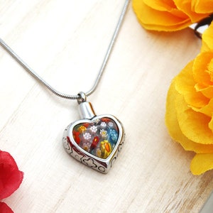 May include: A heart-shaped pendant necklace with a silver chain. The pendant is stainless steel with a decorative floral design and a glass front. The glass front reveals a colorful floral pattern. Red and yellow flowers are in the background.