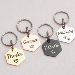 May include: Four personalized pet ID tags with key rings. The tags are hexagonal and made of metal. The tags are gold, silver, and black. The tags are engraved with the names "Phoebe", "Gemma", "Zeus", and "Mickey". Each tag has a small symbol engraved on it, such as a paw print, a heart, a fish, or a star.