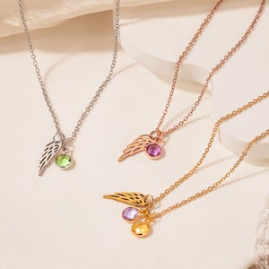 May include: Three necklaces with wing pendants and gemstone accents. One necklace is silver with a green gemstone, another is rose gold with a pink gemstone, and the third is gold with purple and yellow gemstones.