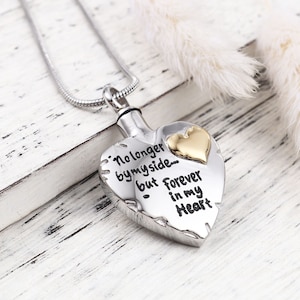 May include: Silver heart-shaped cremation necklace with a gold heart charm. The necklace has an inscription that reads "No longer by my side but forever in my heart."