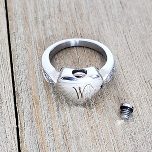 May include: A silver ring with a heart-shaped design and a small screw on the side. The ring is engraved with the letter "W".