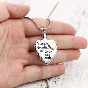May include: Silver heart-shaped cremation necklace with a gold chain. The pendant has an inscription that reads "No longer by my side... but forever in my heart".