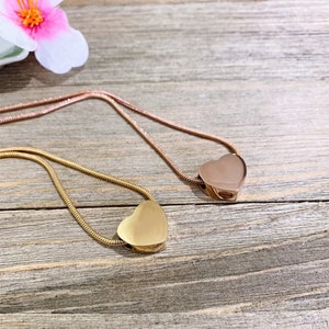 Puede incluir: Dos collares de cremaci&oacute;n en forma de coraz&oacute;n de oro sobre una superficie de madera. Un collar es de oro rosa y el otro es de oro amarillo.