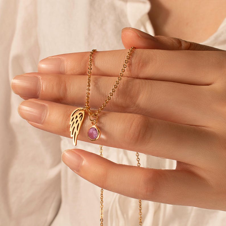 May include: A gold-colored necklace featuring a delicate wing charm and a small, round, pink gemstone pendant. The necklace is held in a hand, showcasing its intricate design and the warm tones of the metal and stone.