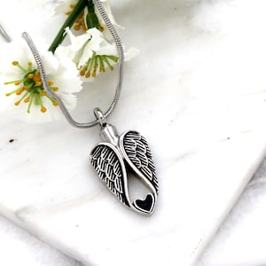 Puede incluir: Collar de cremaci&oacute;n de alas de &aacute;ngel de plata con un coraz&oacute;n negro en el centro. El collar est&aacute; sobre una superficie blanca con flores blancas.
