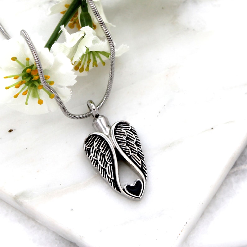 Puede incluir: Collar de cremaci&oacute;n de alas de &aacute;ngel de plata con un coraz&oacute;n negro en el centro. El collar est&aacute; sobre una superficie blanca con flores blancas.