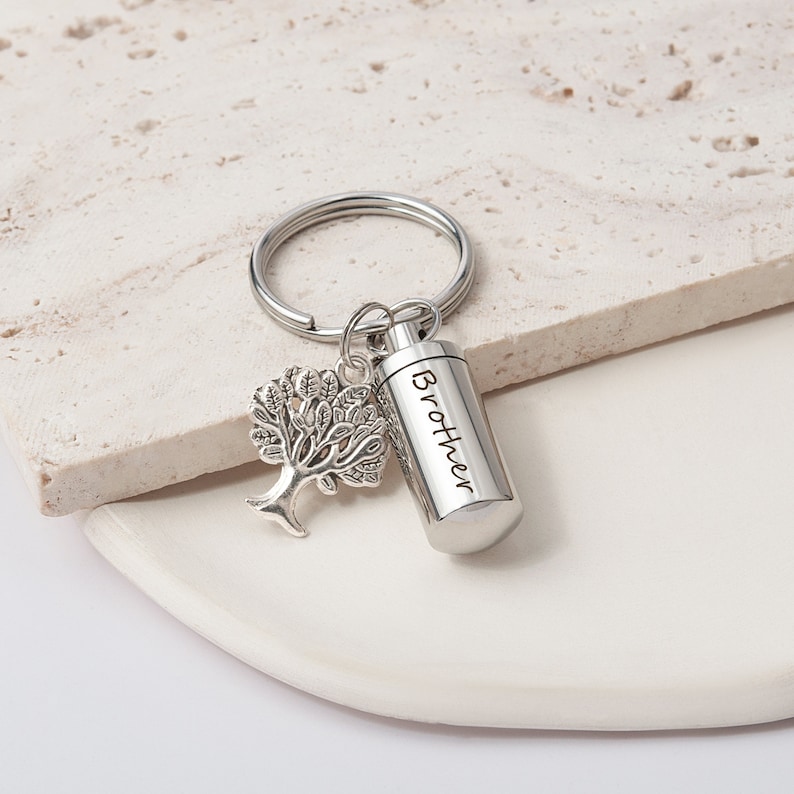 May include: Silver keychain with a small tree charm and a cylinder-shaped cremation urn engraved with the word "Brother".
