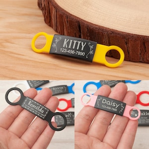 May include: A set of five personalized pet ID tags with black engraved text on a silver background. The tags are attached to colorful silicone rings. The tags feature the names "Romilda", "Kitty", "Daisy", "Bonnie", and "Bo" with phone numbers.