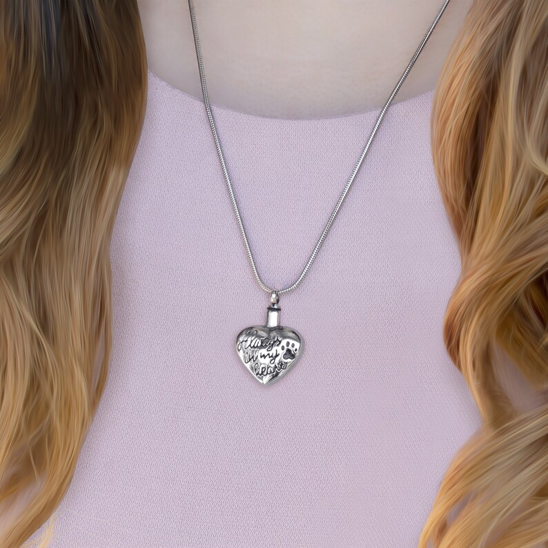 May include: A silver heart-shaped cremation necklace with the inscription "Always in my heart" and a paw print. The necklace is on a silver chain.