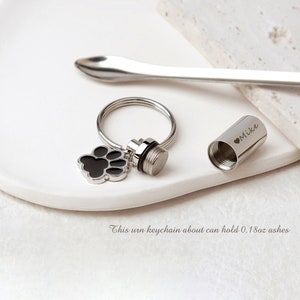May include: Silver keychain with a black paw print charm and a small silver urn. The urn has a screw top and is engraved with "Mike" and a heart. This urn keychain can hold about 0.18 oz of ashes.