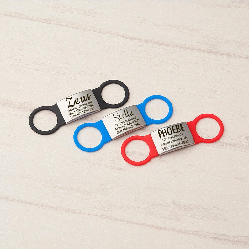 May include: Three rectangular metal pet ID tags with silicone bands in black, blue, and red. Each tag features two circular loops on the sides and is engraved with names and contact information, including Zeus, Stella, and Phoebe with phone numbers.