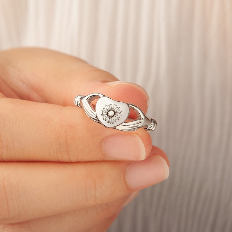 May include: Silver ring with a heart-shaped design featuring a delicate flower in the centre.