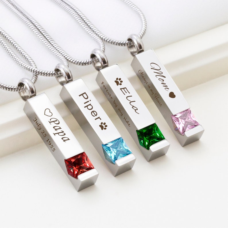 May include: Four silver bar necklaces with different colored gemstones and engraved messages. The necklaces are engraved with the names "Mom", "Ella", "Piper", and "Papa". The messages on the necklaces are "Always in my Heart", "Forever by my side", and "July 25 1955".