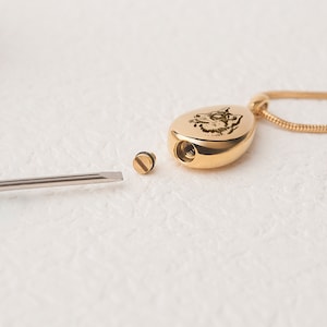 May include: Gold teardrop-shaped cremation pendant with a wolf engraved on the front. The pendant is open and the lid is off, revealing the inside of the pendant. A small screwdriver is also shown.