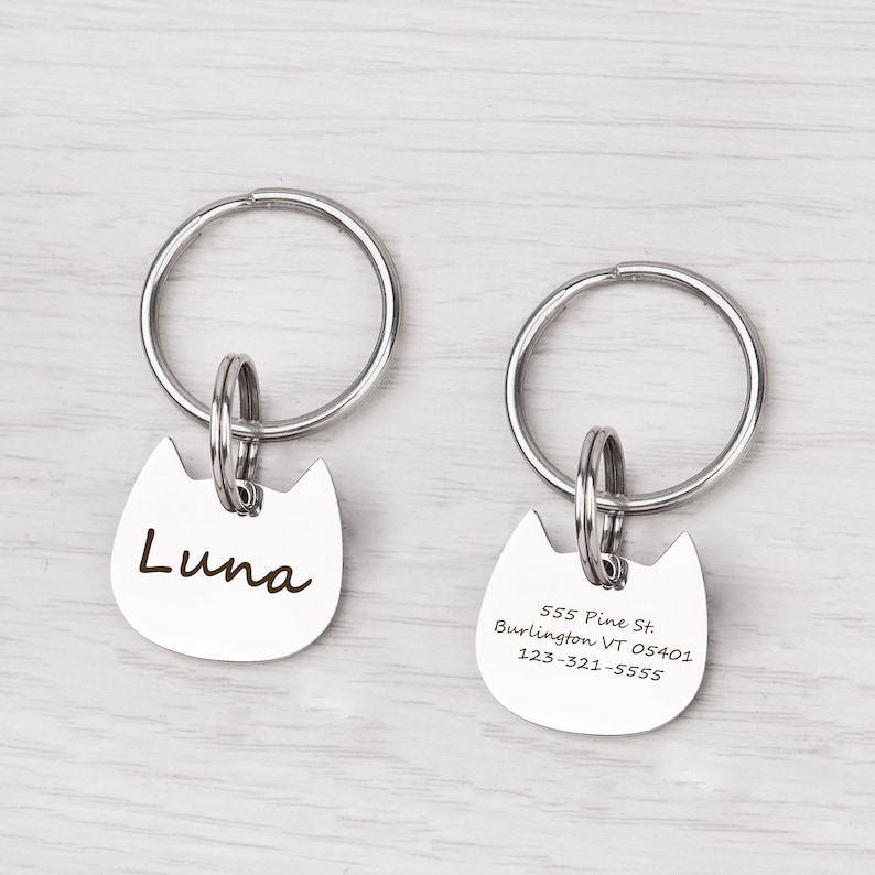 Puede incluir: Dos placas de identificaci&oacute;n para mascotas de metal plateado con forma de gato, unidas a anillas circulares. La placa izquierda tiene grabado el nombre Luna en letra cursiva. La derecha muestra la direcci&oacute;n 555 Pine St. Burlington VT 05401 y el n&uacute;mero 123-321-5555.