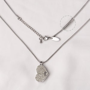 Puede incluir: Un collar de plata con un colgante en forma de coraz&oacute;n que presenta un dise&ntilde;o de huella digital. El collar tiene una cadena con un cierre de langosta y una cadena extensora.