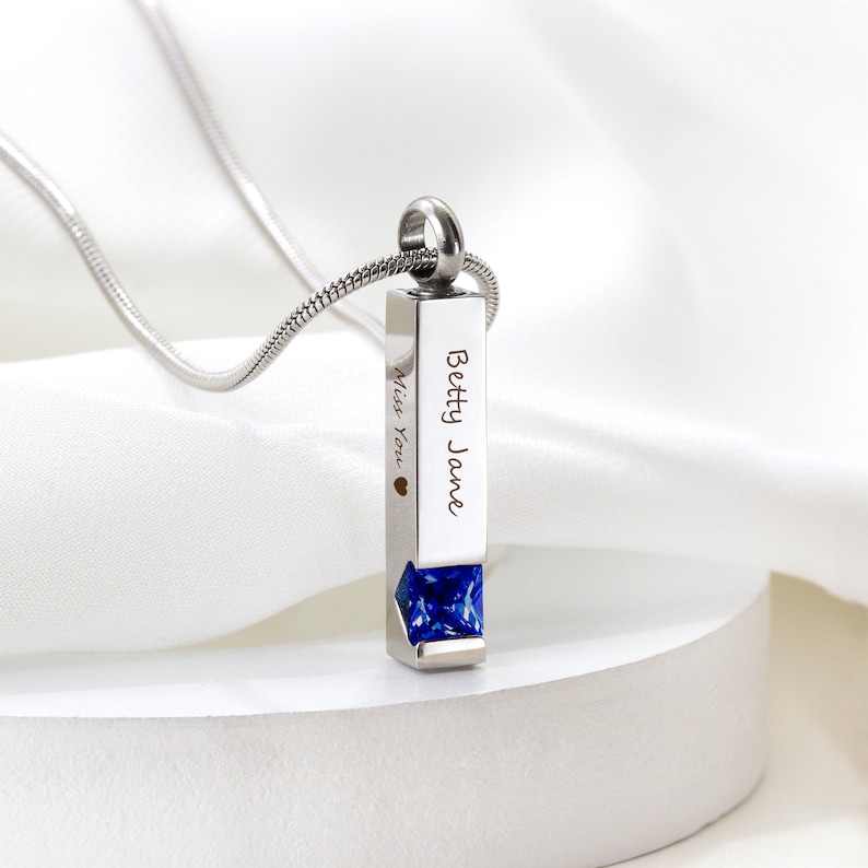 May include: A silver necklace with a rectangular pendant that has the name "Betty Jane" engraved on it. The pendant has a blue gemstone at the bottom and the words "Miss You" engraved on the side.