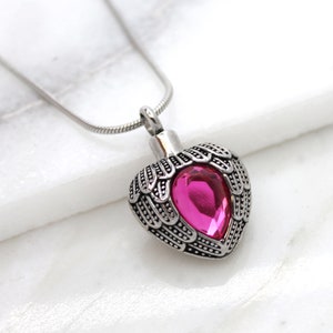 May include: A silver heart-shaped cremation necklace with angel wings and a pink crystal. The necklace is on a silver chain.