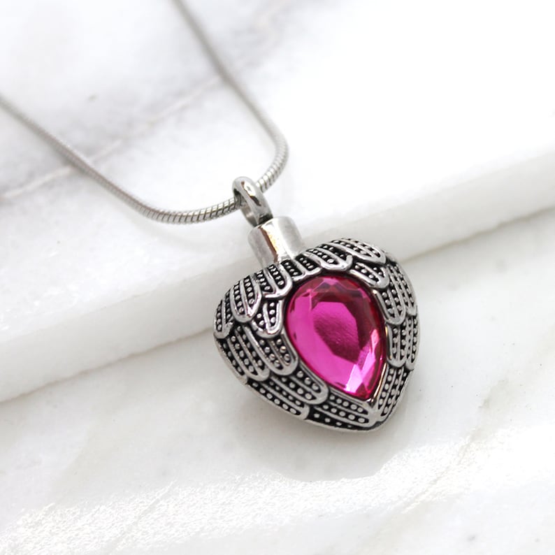 May include: A silver heart-shaped cremation necklace with angel wings and a pink crystal. The necklace is on a silver chain.