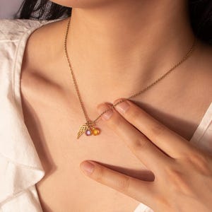 May include: A gold-colored necklace with a delicate chain. The pendant features a small wing charm, a purple gemstone, and a yellow gemstone. The necklace is worn against a white shirt.