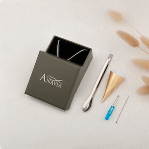 May include: A silver necklace in a green box with the word "Anavia" on it. The box is next to a silver tool, a gold paper cone, a blue screwdriver, and a silver needle.