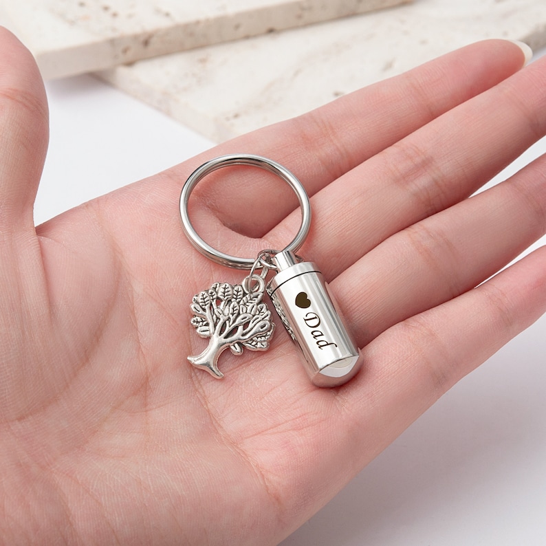 May include: Silver keychain with a small tree charm and a cylindrical cremation urn engraved with "Dad" and a heart.