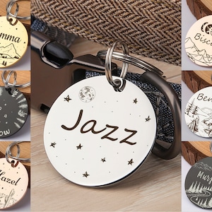 Op de afbeelding: Een assortiment gepersonaliseerde hondenpenningen met verschillende ontwerpen. De penningen zijn gegraveerd met namen zoals "Gemma", "Willow", "Hazel", "Jazz", "Biscuit", "Benny" en "Murphy". Elke penning heeft een uniek ontwerp met een berg-, bos- of hemelthema.