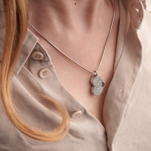 Puede incluir: Un collar de cadena de plata con un colgante de coraz&oacute;n doble. El colgante est&aacute; grabado con un dise&ntilde;o de huella digital.