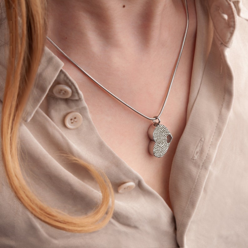 Puede incluir: Un collar de cadena de plata con un colgante de coraz&oacute;n doble. El colgante est&aacute; grabado con un dise&ntilde;o de huella digital.