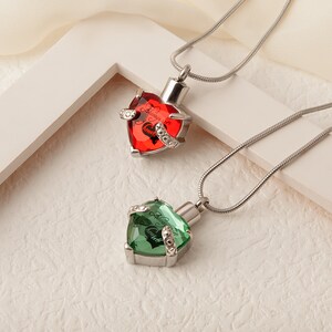 May include: Two silver heart-shaped cremation necklaces with a chain. The top necklace has a red crystal with the inscription "Always in my heart". The bottom necklace has a green crystal with the inscription "Always in my heart".