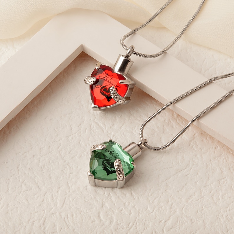 May include: Two silver heart-shaped cremation necklaces with a chain. The top necklace has a red crystal with the inscription "Always in my heart". The bottom necklace has a green crystal with the inscription "Always in my heart".