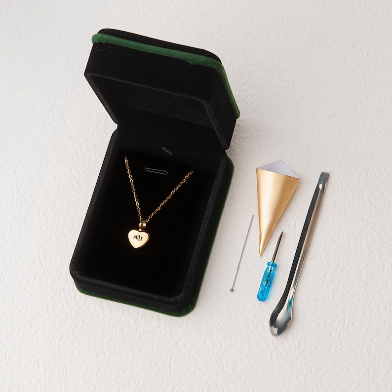 May include: A gold heart-shaped pendant necklace with the initials "MLV" engraved on it, comes in a black velvet box with a gold funnel, a small screwdriver, a spoon-shaped tool, and a small pin.