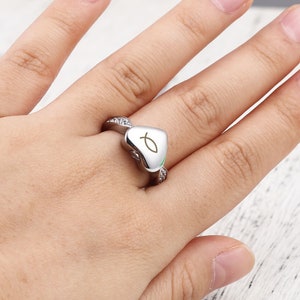 May include: A silver heart-shaped ring with a small fish symbol engraved on the front. The ring is set with small clear stones around the band.
