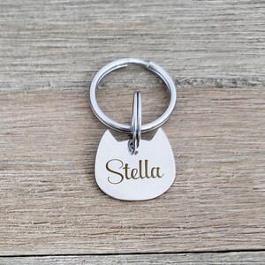 Puede incluir: Una placa de identificaci&oacute;n para mascotas de metal plateado con forma de cabeza de gato y orejas puntiagudas. La placa tiene el nombre Stella grabado en letra cursiva y est&aacute; sujeta a una anilla met&aacute;lica circular para el collar.