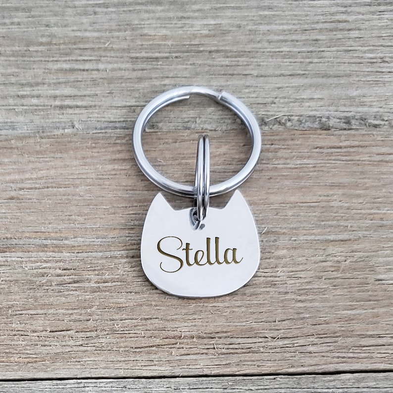 Puede incluir: Una placa de identificaci&oacute;n para mascotas de metal plateado con forma de cabeza de gato y orejas puntiagudas. La placa tiene el nombre Stella grabado en letra cursiva y est&aacute; sujeta a una anilla met&aacute;lica circular para el collar.