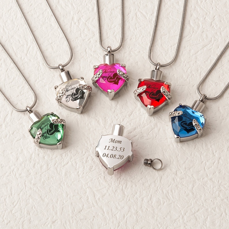 May include: Five silver heart-shaped cremation necklaces with different colored crystals. The necklaces have the inscription "Always in my heart" on the front. One necklace has a plain silver heart with the inscription "Mom 11.23.53 04.08.20".