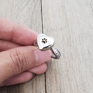 May include: A silver heart-shaped ring with a small paw print engraved on the front. The ring has a band of small clear stones.
