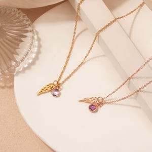 May include: Two gold-colored necklaces with wing pendants and small, round, purple gemstones. The necklaces are displayed on a white surface, with a clear glass dish in the background. The chains are delicate and the wings have detailed feather designs.