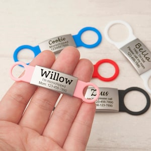 May include: A rectangular metal pet ID tag with a pink silicone band featuring the name Willow, the text I'm microchipped, and the phone number 123-456-7890. Additional tags in blue, white, red, and black are visible with various names and contact details.