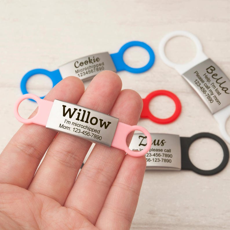 May include: A rectangular metal pet ID tag with a pink silicone band featuring the name Willow, the text I'm microchipped, and the phone number 123-456-7890. Additional tags in blue, white, red, and black are visible with various names and contact details.